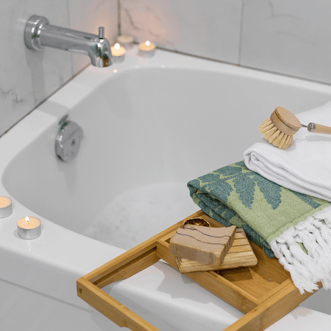 Green leaf Turkish towel and white Turkish towel folded on top of a bath tray within a candle lit bath tub.