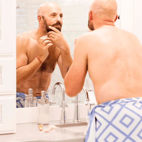 A bald man with a beard, shirtless looks at himself in a mirror wearing only a Turkish towel