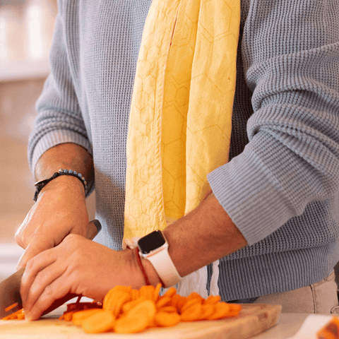 A mans hands chop orange carrots. He wears a watch. A yellow honeycomb Turkish cotton tea towel is draped over a shoulder