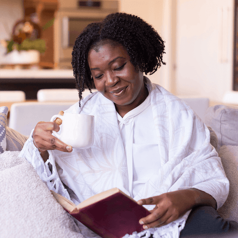 Woman of colour draped in a white Turkish towel reading a book while holding a mug.