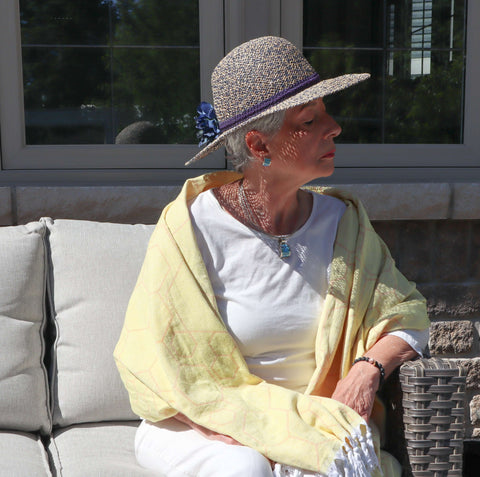 Elderly woman sits outside with a hat wearing a yellow orange honeycomb Turkish towel over her shoulders