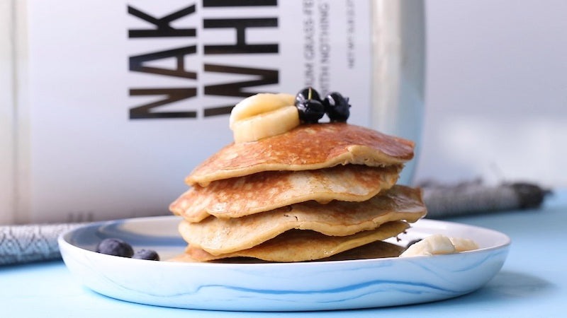 Stack of pancakes with blueberries and banana slices on top, with a tub of Naked Whey in the background