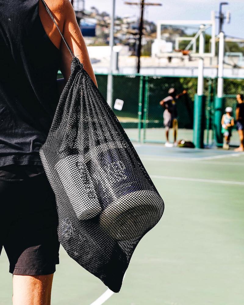 Man walking on a tennis court, carrying a mesh bag with a tub of Naked Mass and a Naked Nutrition shaker bottle
