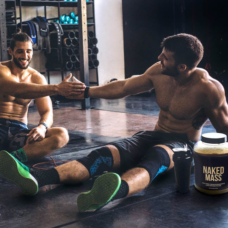 Two men sitting down and resting after a workout in the gym, a tub of Naked Mass is next to them