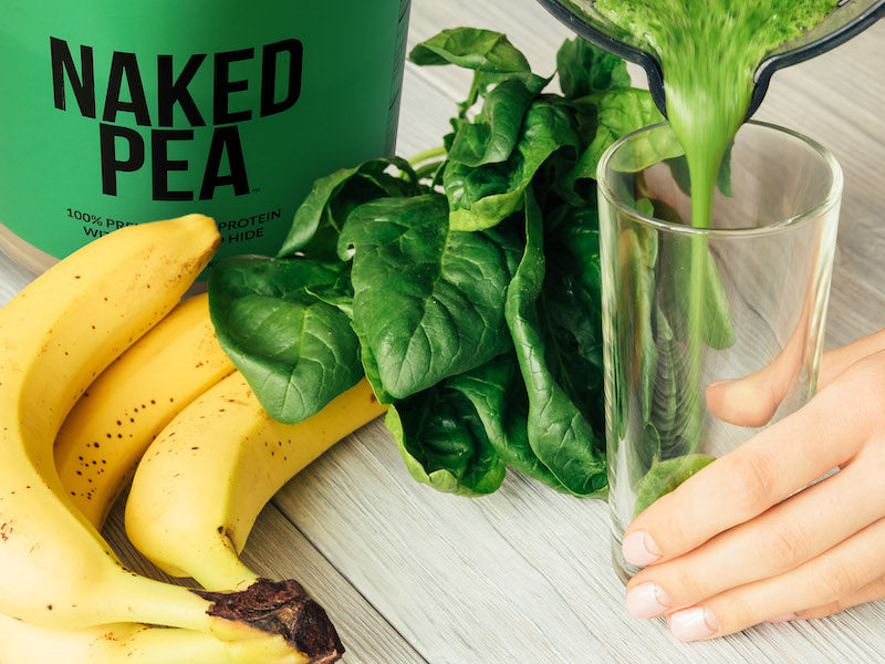 Naked Pea product image with a protein shake being poured into a glass next to the product, spinach leaves, and bananas
