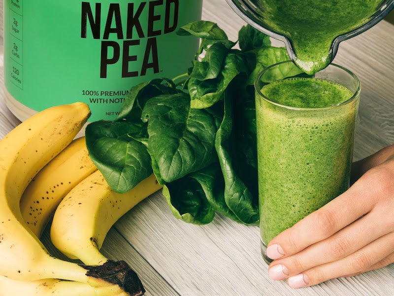 Naked Pea product image with a tub of the product next to a green pea protein shake poured into a glass with spinach leaves and banana