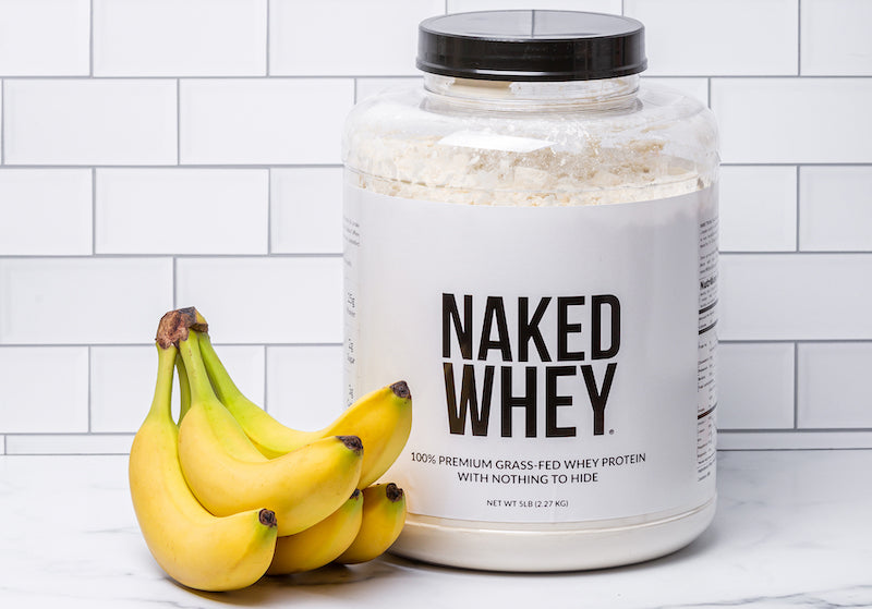 Tub of Naked Whey next to a bunch of bananas in front of a white wall