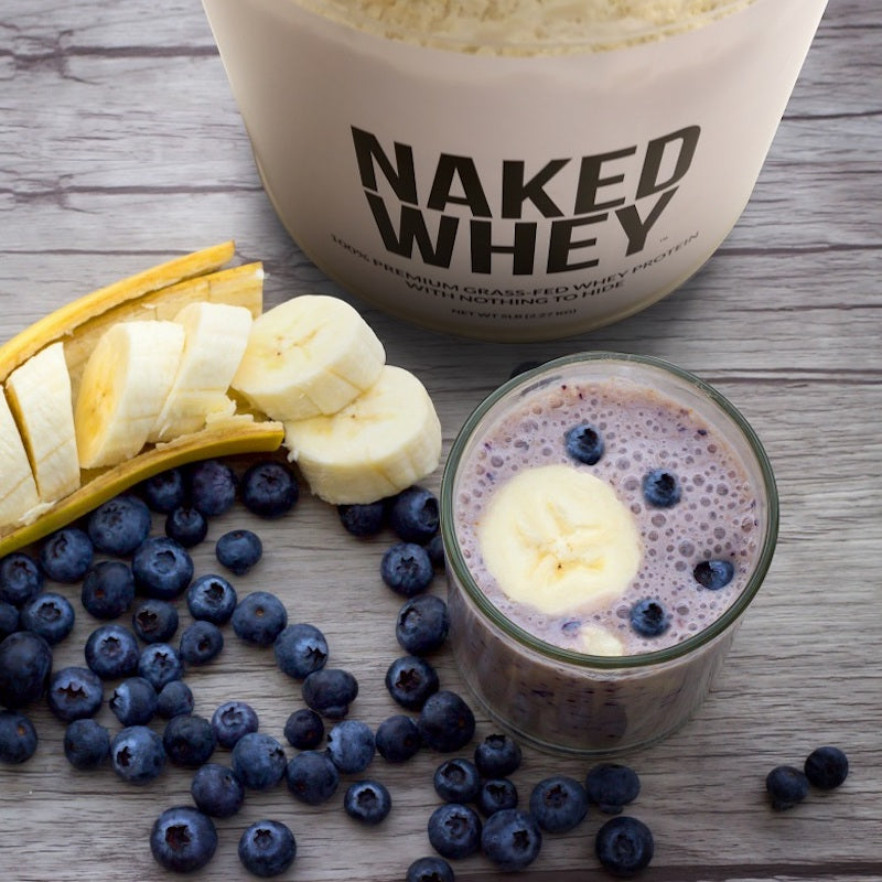 Tub of Naked Whey protein powder next to a blueberry and banana protein shake with the ingredients next to it