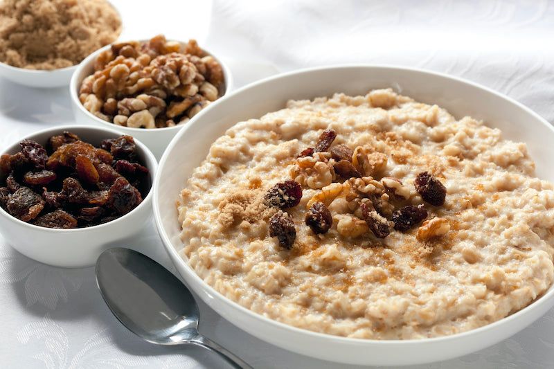 Cooked oatmeal in a bowl, next to smaller bowls of raisins, nuts, and brown sugar