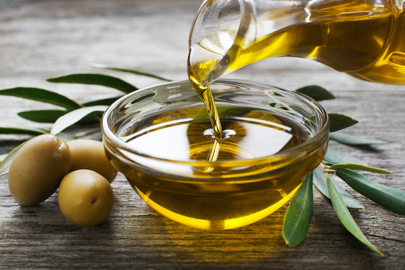 Olive oil being poured into a clear bowl, with raw olives to the side