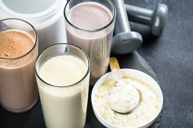 Three different flavored protein shakes next to a tub of protein powder, a dumbbell, and a scoop of protein powder