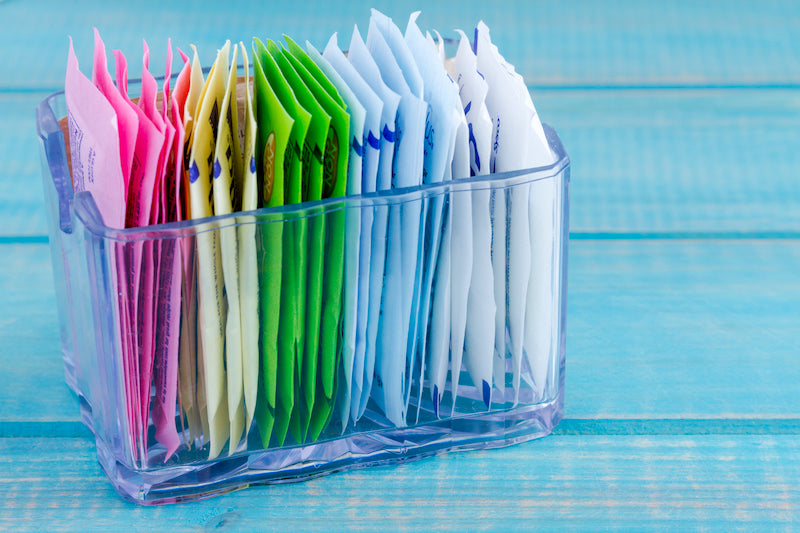 Packets of artificial sweeteners in a glass holder on a blue wooden table