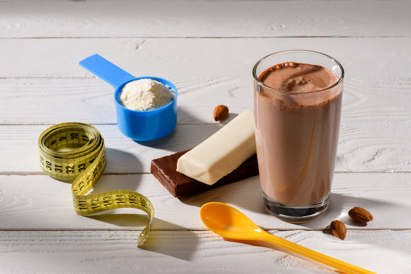 Weight gainer protein shake next to a tape measure, a plastic spoon, two chocolate bars, and a scoop of protein powder