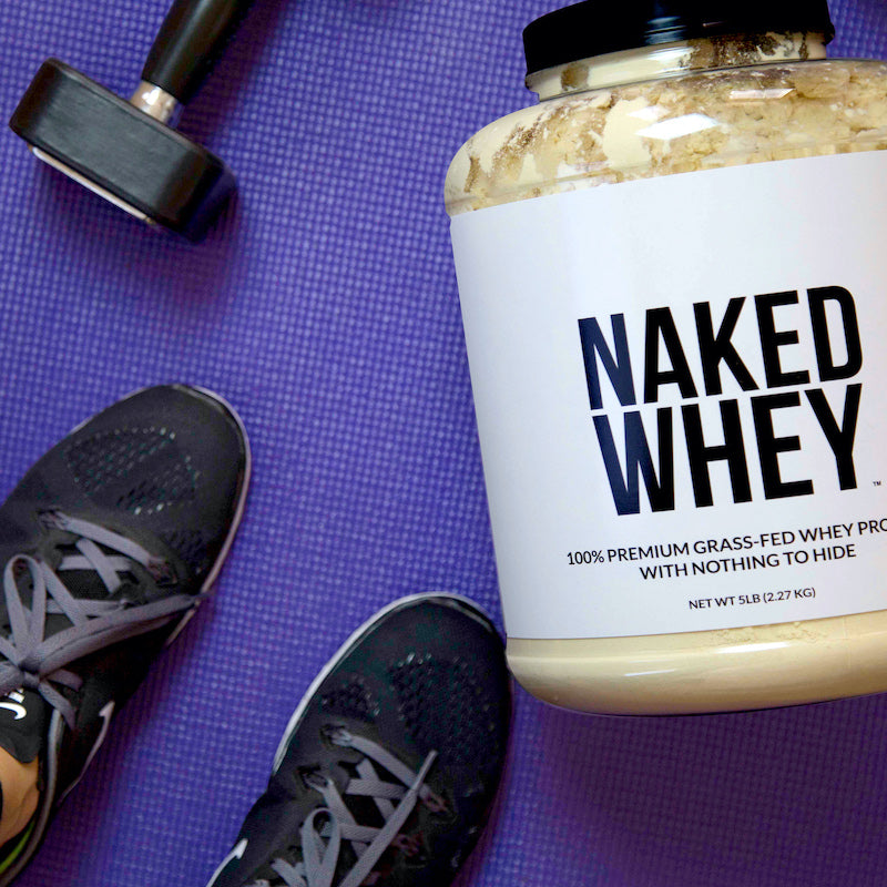 Tub of Unflavored Naked Whey on a gym mat next to a pair of gym shoes and a weight