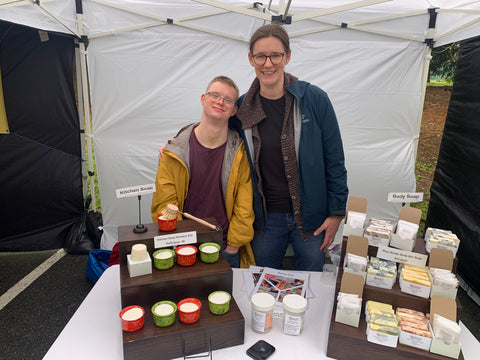 man in yellow jacket and woman in teal jacket stand behind table of soap products