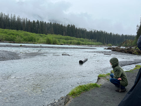 a teenager squats next to the Hoh River. A wet scene, there is driftwood, rocks, and aquamarine water in the foreground.  The midground is multiple shades of green, topped with a thick layer of fog that hides the mountains beyond.