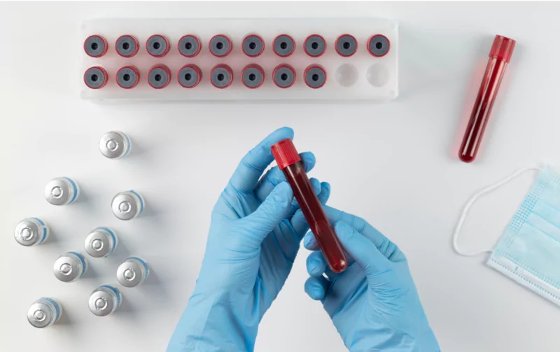 Hands in gloves holding a blood test tube with vials and a face mask on a white surface.