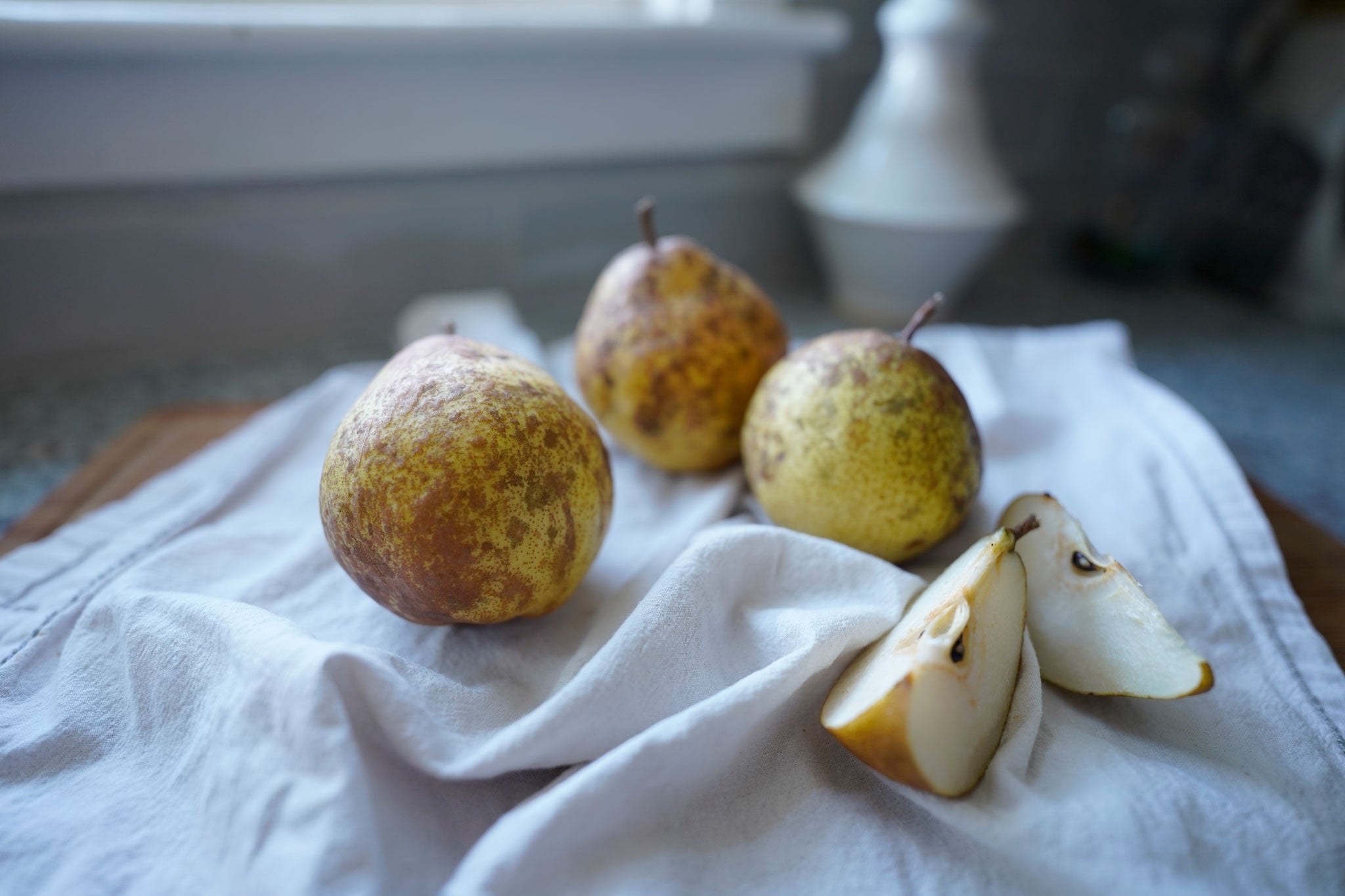Pears and pear slices on white dish towel and wood cutting board on kitchen counter