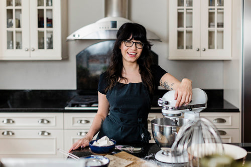 marshmallow girl with bangs, glasses, and long brown hair stands smiling in her kitchen