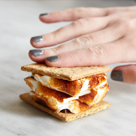 hand squishing a s'more with handcrafted marshmallows on marble table