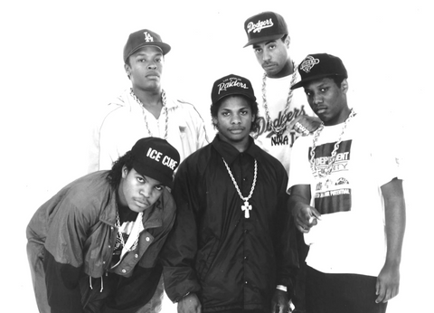Black and white group portrait of NWA members wearing Los Angeles Dodgers and Raiders caps and sporting chain necklaces, emblematic of their influence in the gangsta rap genre.