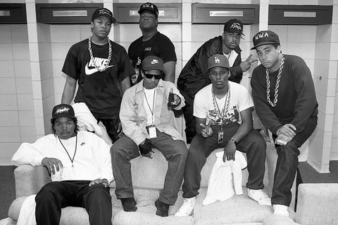 Black and white photo of NWA members posed in a backstage area during their 1989 tour, sporting casual sportswear and accessories that showcase their gangsta rap image.