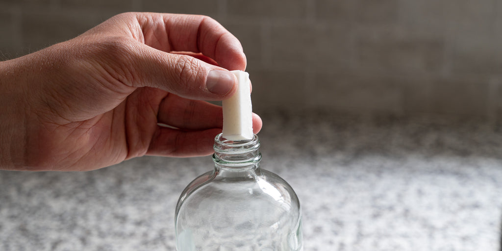 A hand dropping an all-purpose cleaner tablet into a glass spray bottle