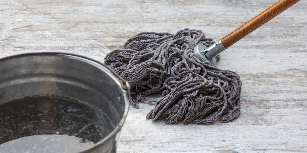 A wood-and-metal mop and metal bucket against a faux-wood laminate floor