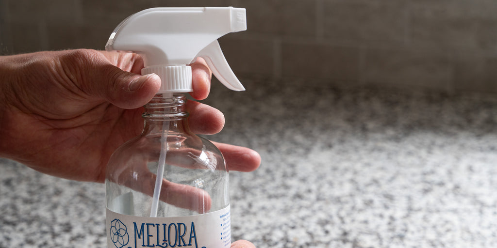 A hand unscrewing the sprayer attachment from a Meliora all-purpose cleaner bottle