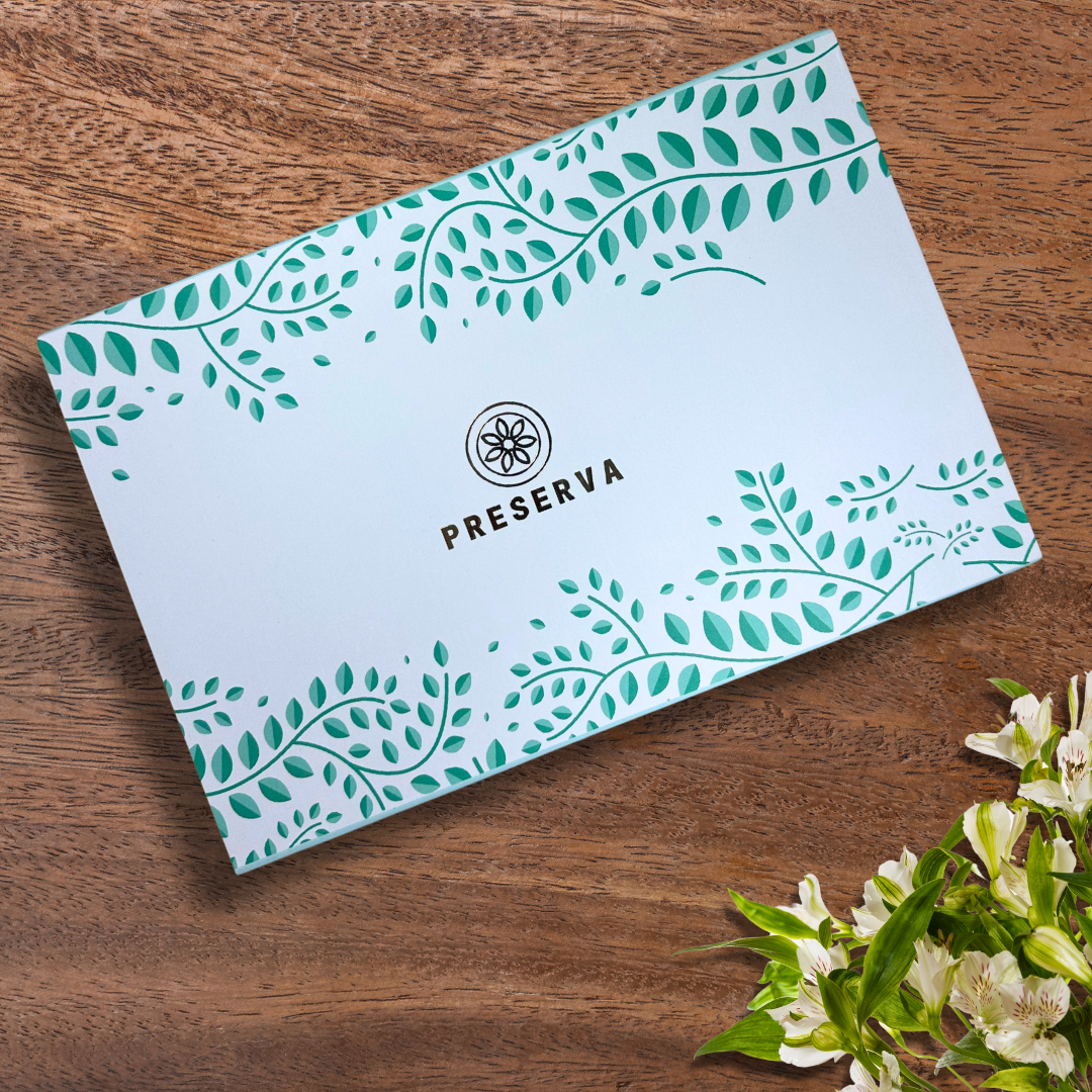Green Preserva Wellness Gift Box with leaf pattern on it next to white flowers on a wooden table