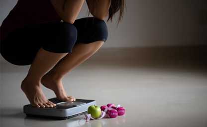A woman stressed and sitting on a weighing machine next to an apple, measuring tape, and dumbbells