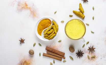 Turmeric milk with raw and powdered turmeric in a white tray surrounded by cinnamon and herbs