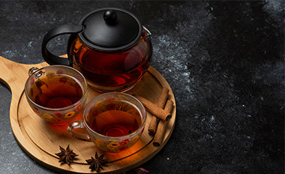 Two cups of black tea with a teapot on a wooden tray next to cinnamon sticks