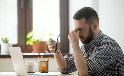 A person rubbing their eyes while working on a laptop