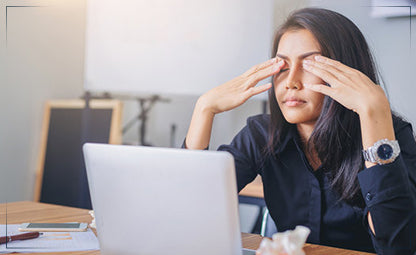 A woman with her hands on her eyes in the office in front of the laptop