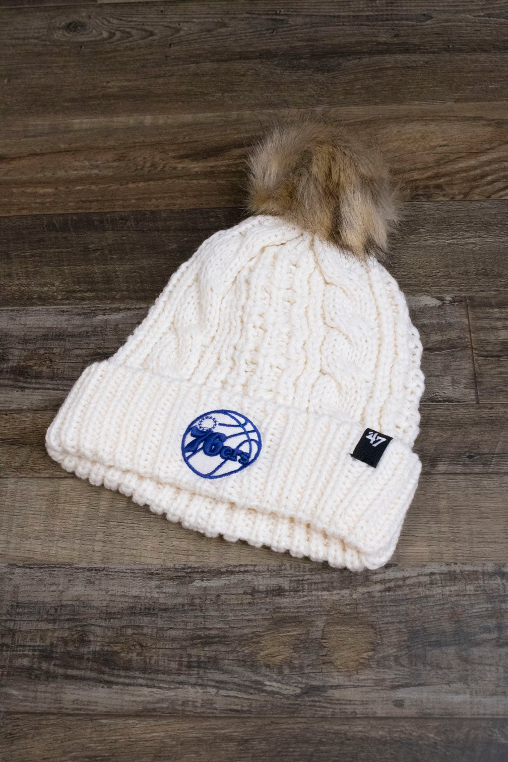 the front of the Philadelphia 76ers Ladies Fur Pom Cream Winter Beanie | Sixers Meeko Faux Fox Fur White Pompom Beanie has a tonal Sixers blue and white logo