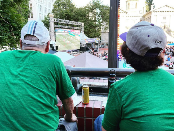 An over-the-shoulder photo of two people watching the Women's World Cup over a huge screen in downtown Portland