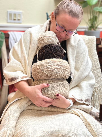 Caucasian female with glasses and hair pulled back into a braid, sits with a wool blanket on her lap, a wool shawl around her shoulders, and lovingly cradles wheels of wool yarn stacked in her arms.