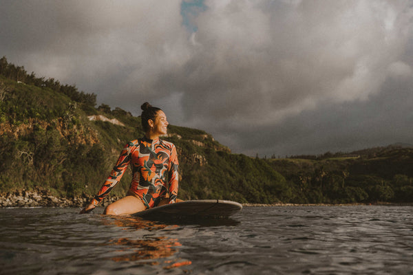 Female surfer sitting on her surfboar in the ocean, smiling and wearing a long sleeve spring surfsuit by SunDaze Surf