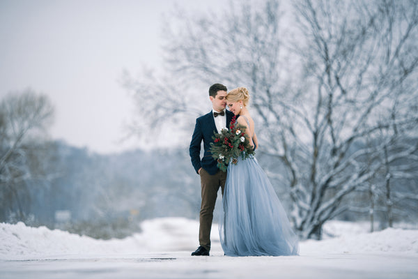 Outdoor winter wedding bride and groom photo groom in suit and bride is dressed in periwinkle blue wedding gown holding a bouquet of red and white roses