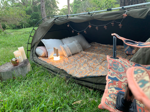 picture of a camping swag with a boho sleeping bag and camping chair. The sway has extra pillows and cushions with a carry lamp. 