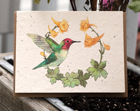 A photo of a Bower Studio Seed card featuring an illustration of a green and red Anna’s Hummingbird surrounded by yellow Larkspur. The card is sitting outdoors on a wooden planter box.