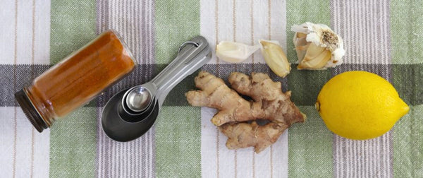 A photo of a bottle of cayenne pepper, measuring spoons, ginger root, garlic, and a lemon on a green plaid cloth