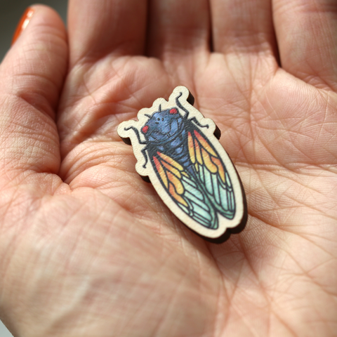 a photo of our original wooden cicada pin in the palm of a pale hand