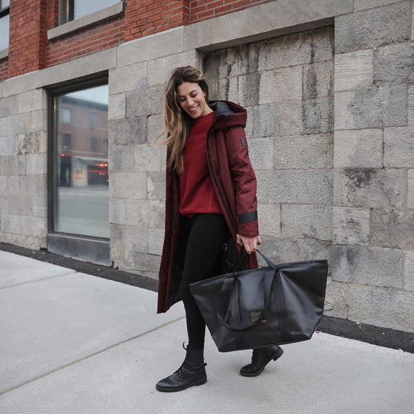A female model wearing a red Bedi coat and carrying a black dufflepack bag