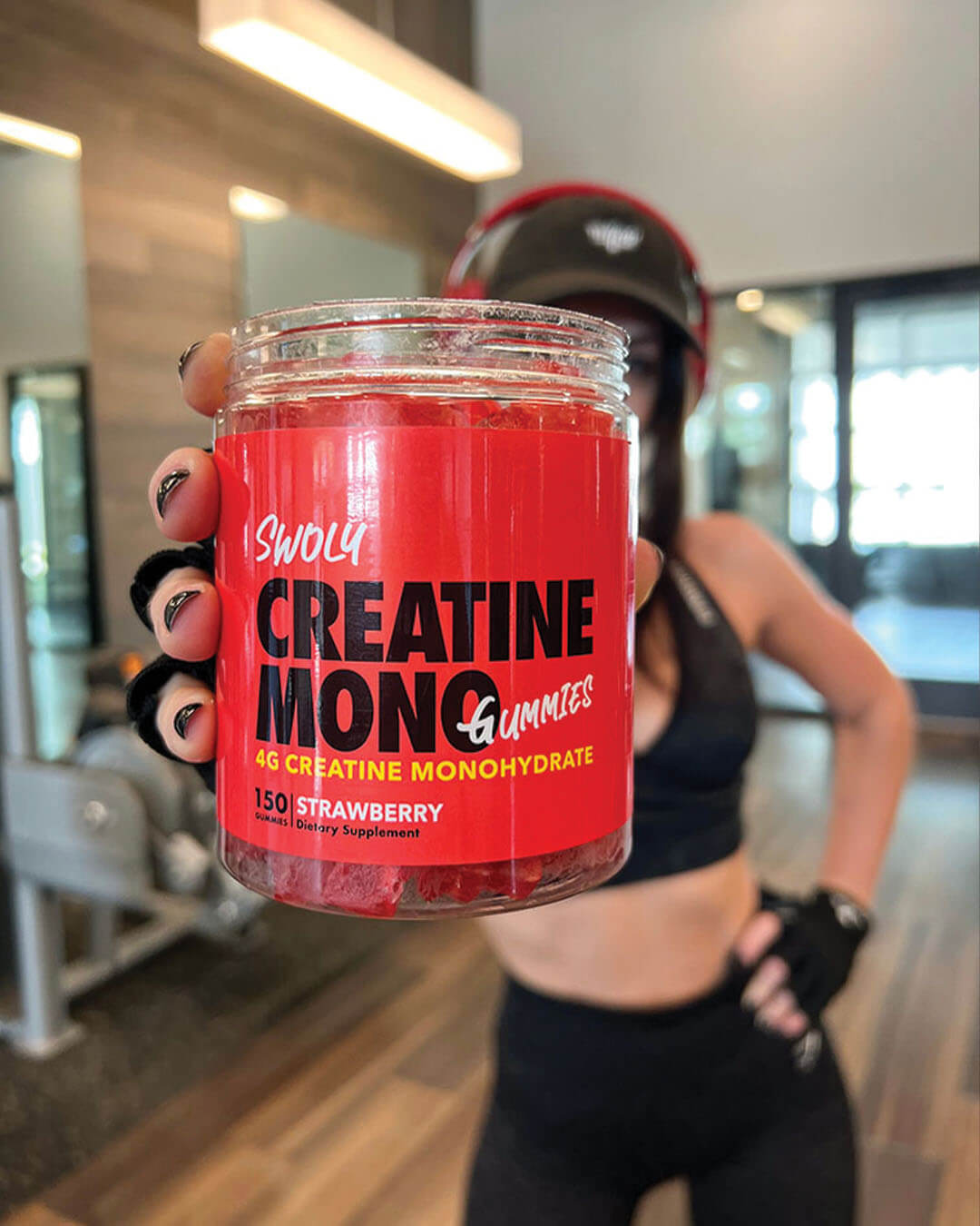 athletic woman holding creatine gummies bottle with gym setting in the background, swoly, gummy supplement 