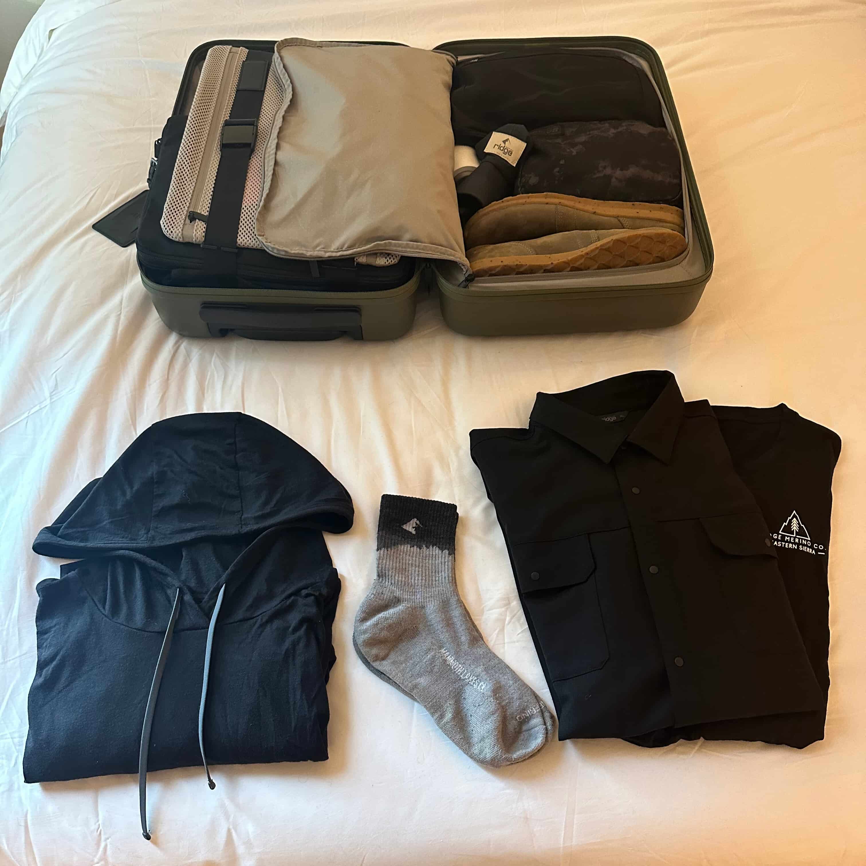 overhead view of a small, well-packed suitcase for travel with the Ridge Solstice Sun Hoodie, High Country Button Down, Minaret Socks and Natural Tee laying below it