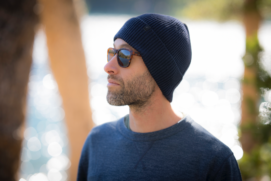 a man wears a Ridge Merino wool beanie and sunglasses standing next to an alpine lake