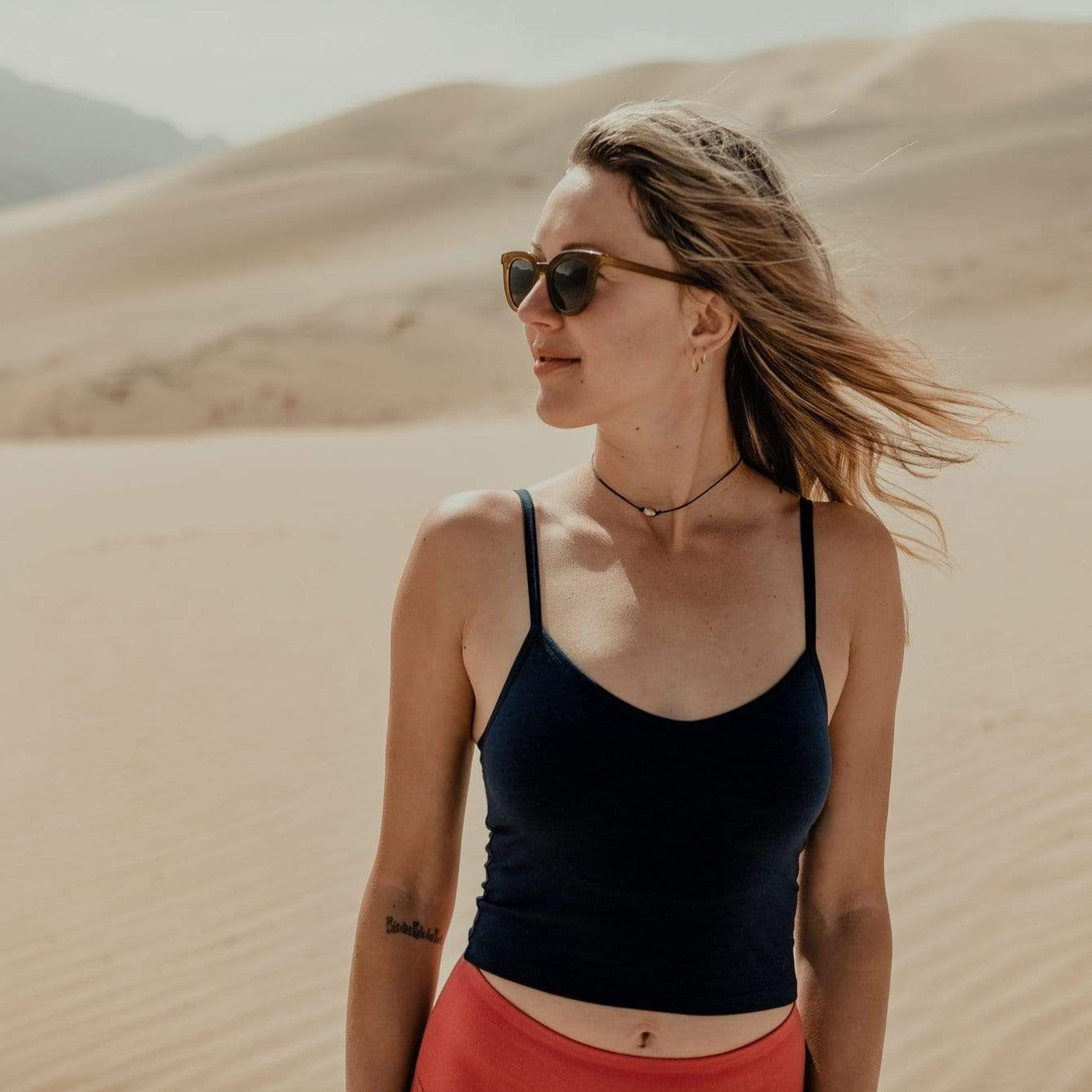 A woman smiles wearing sunglasses and the Merino wool bralette in a sand dune