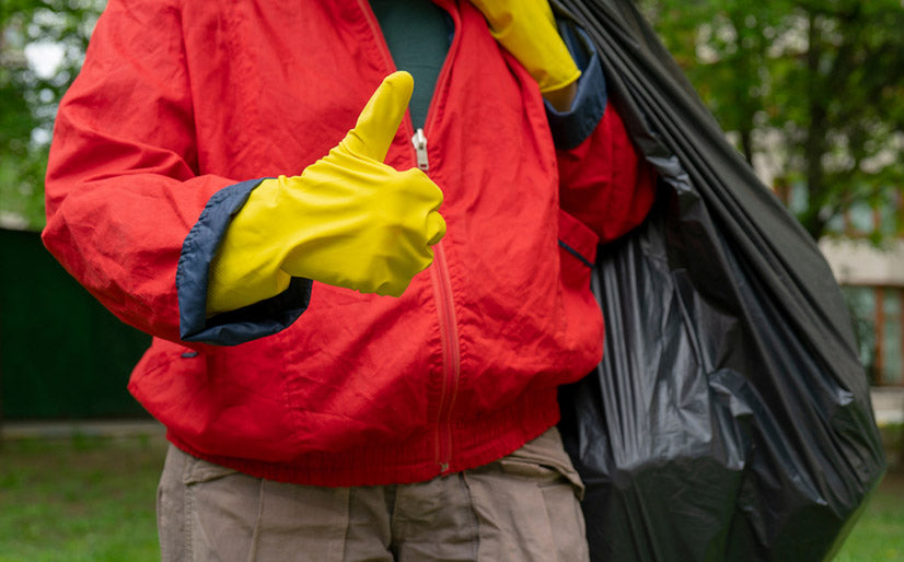 Garbage collection. Man in red coat and yellow gloves showing thumb up, carries a big black bag with trash.